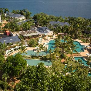 Vista aérea de un complejo turístico con piscinas, palmeras y edificios frente al mar.
