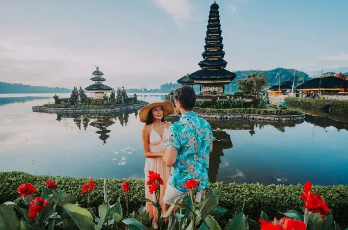 Pareja frente a templos balineses en un lago con flores rojas.