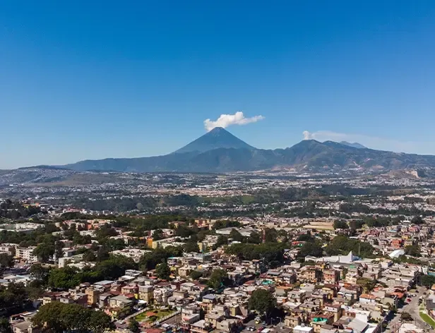 Vista aérea de una ciudad con montañas y volcán humeante al fondo.