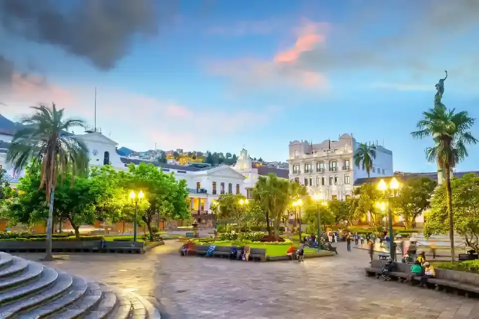 Plaza de ciudad con edificios iluminados, personas y palmeras bajo el cielo crepuscular.