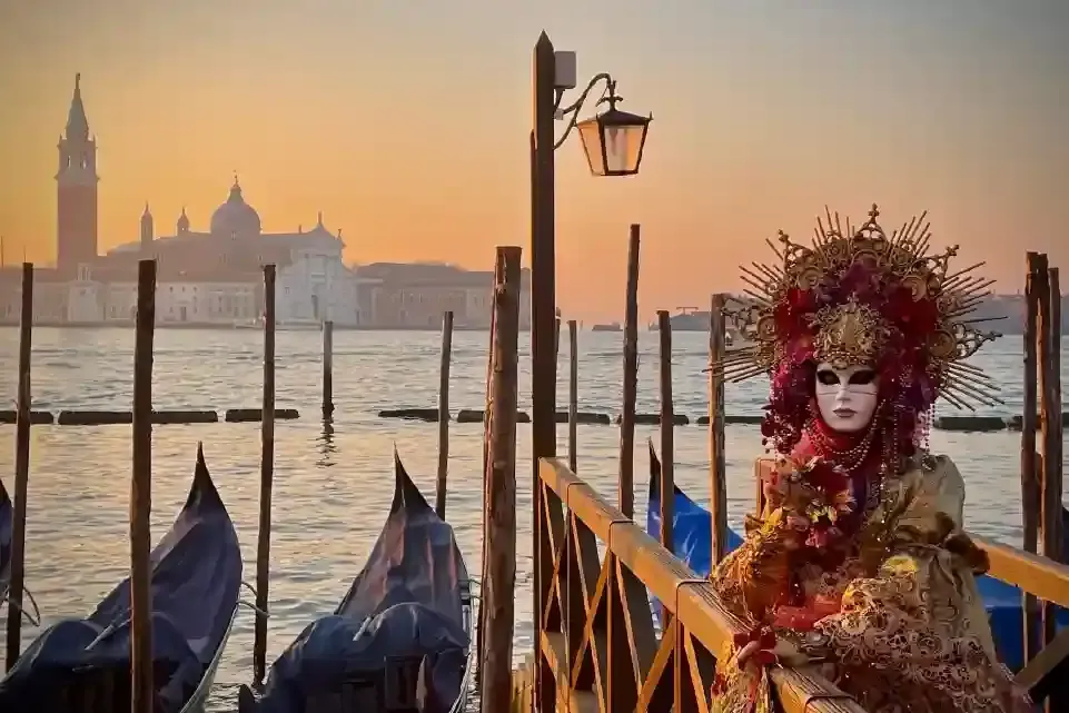 Persona con máscara de carnaval en un muelle con góndolas y Venecia al atardecer.