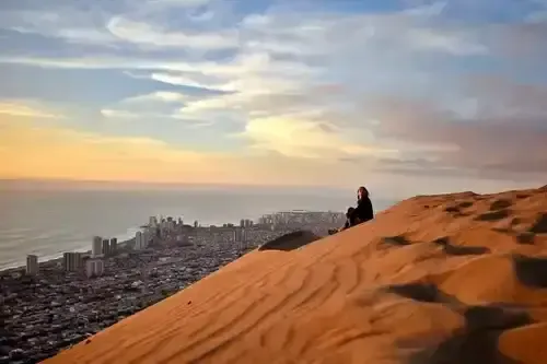 Persona sentada en una duna de arena con una ciudad costera y el mar de fondo.